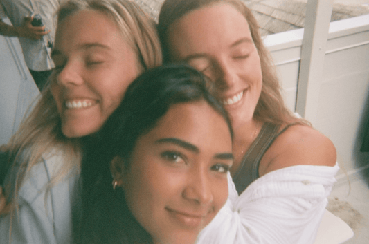 Three girls huddle together, two blonds in the background with their eyes closed and one brunette looking straight at the camera