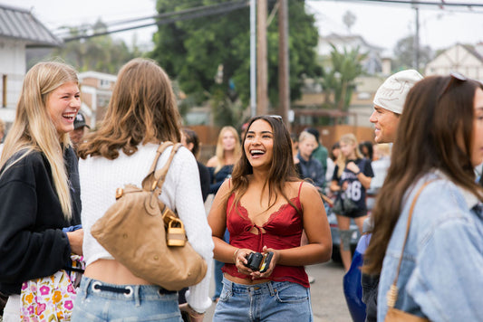 Sienna Mae Gomez, in a red lace tank top, smiles and talks with a group of people outdoors at the Sienna Swim pop-up, with a crowd gathered in the background.