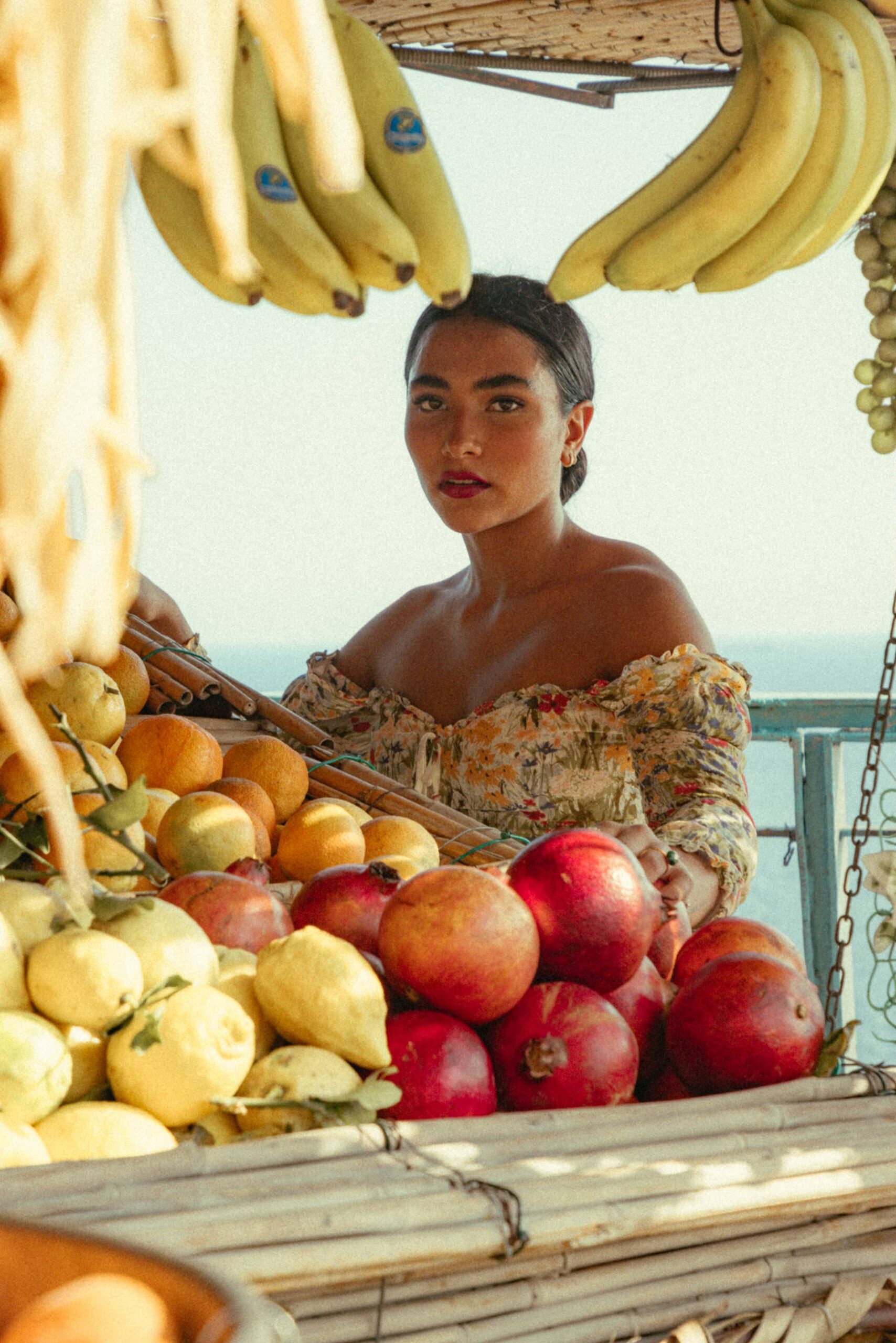 Sienna Mae Gomez surrounded by bananas, oranges, and pomegranates, wearing a yellow floral dress near the sea in Positano