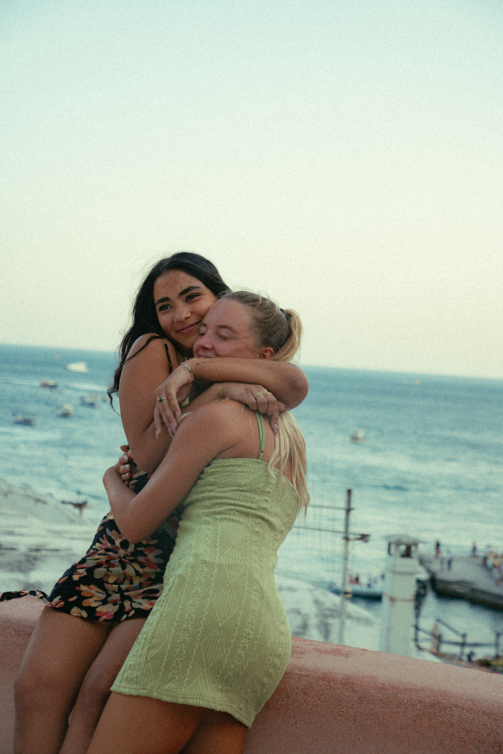 Two girls hugging near the sea, one blonde and one brunette each wearing dresses