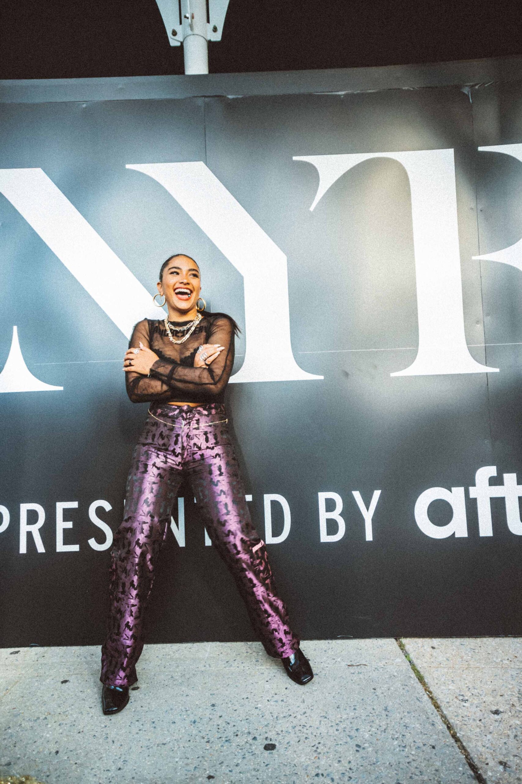 Sienna Mae Gomez in front of NYFW billboard, girl wearing purple pants and laughing