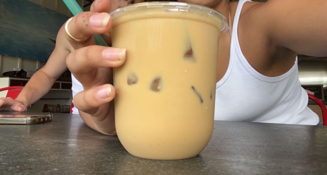 three fingers wrapped around the left hand side of a rounded edge plastic coffee cup, with a few visible ice cubes and filled to the top with caramel colored coffee. Girl in white tank top behind the coffee cup snapping photo.