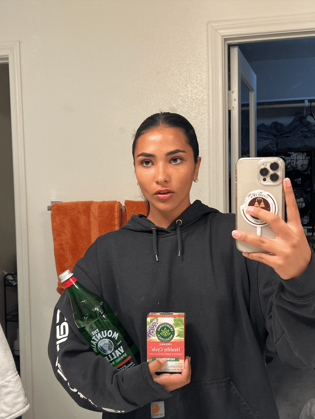 Girl takes mirror selfie with white iPhone 13, wearing a charcoal colored sweatshirt while holding a bottle of mountain valley spring water in her left arm and organic raspberry tea in her left hand