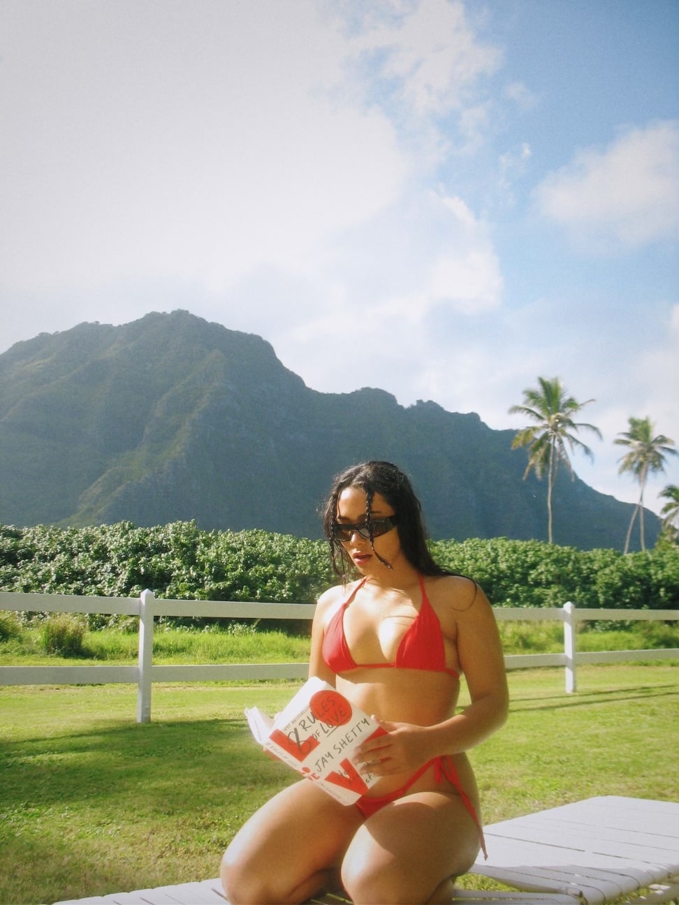 Girl sitting on white lawn chair in red bikini reading book with mountain and palm tree back drop