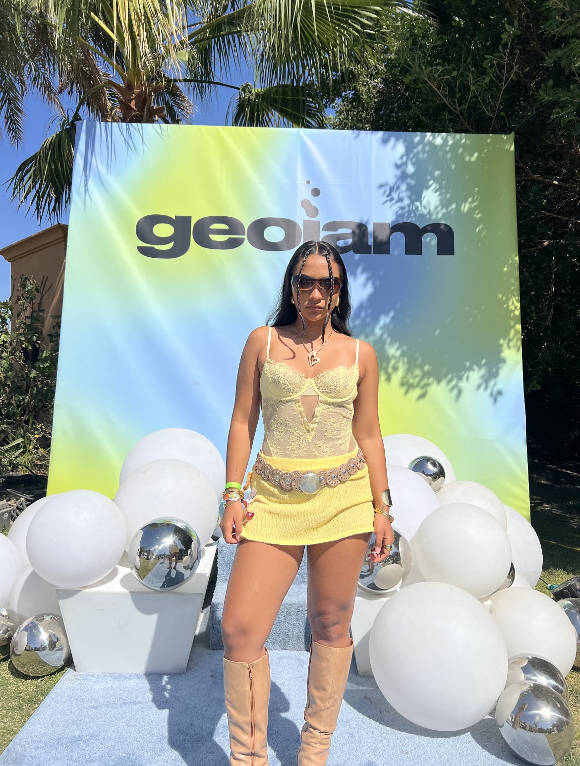 Girl in yellow top, skirt, brown belt, and tan boots posing in front of a Geojam banner