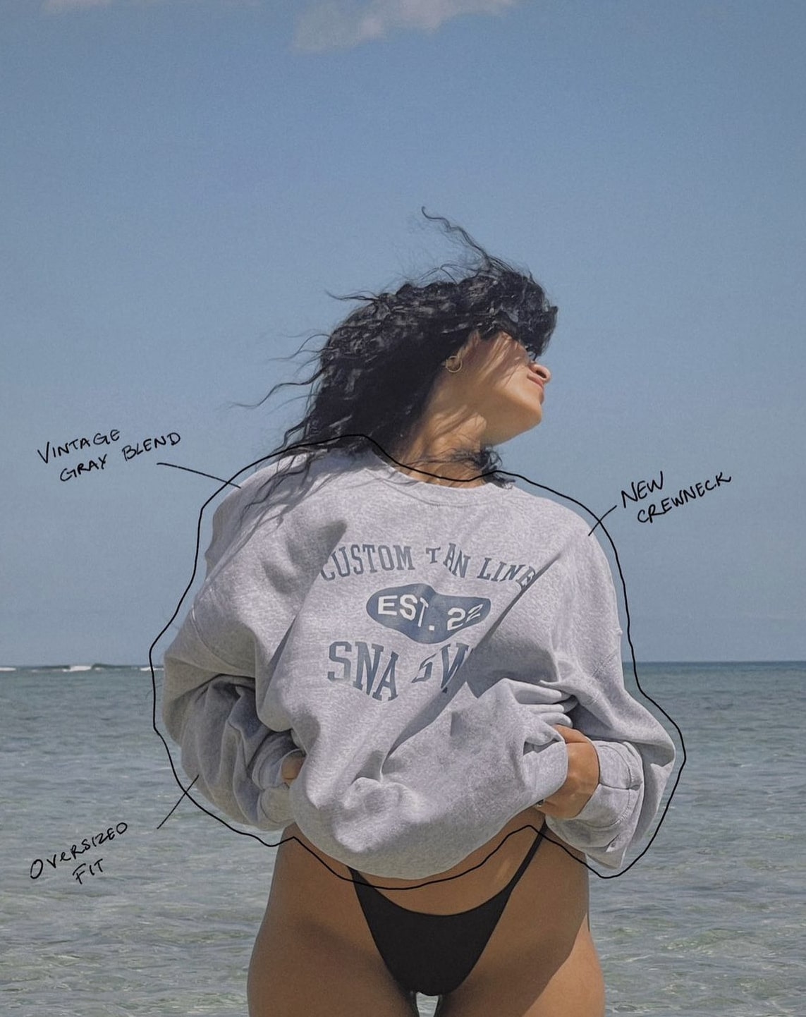 Girl posing in front of ocean wearing a sweatshirt that reads top to bottom left to right, "Custom Tan Lines, Est. 22, SNA SWM"