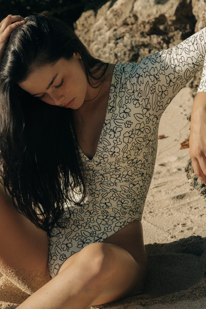 girl sitting in the sand wearing a white, long sleeve surf suit with v neck detail and black hawaiian print flowers