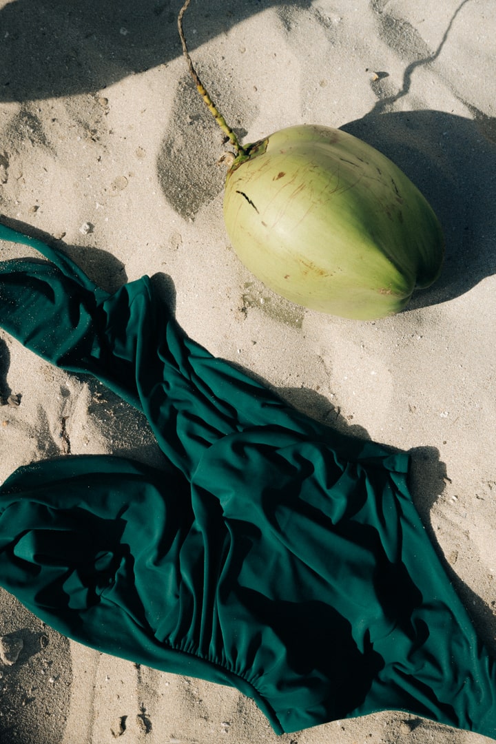 Green one piece bathing suit laying flat in the sand beside a lime green colored coconut