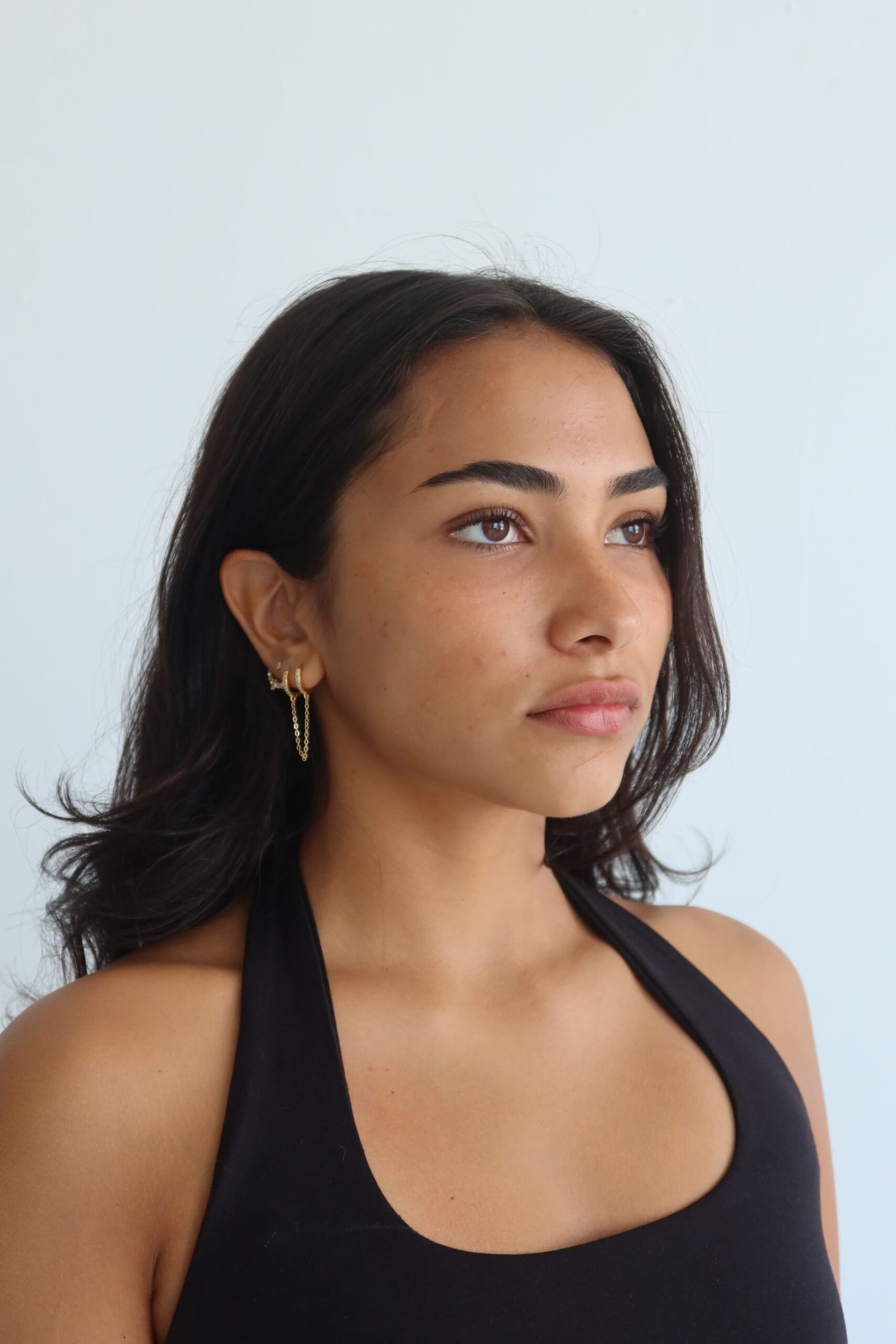 Profile shot of girl looking past the camera against a white backdrop; model wears a black halter top, dangling gold earrings, and light to no makeup. Tan skin, dark brown curly hair.