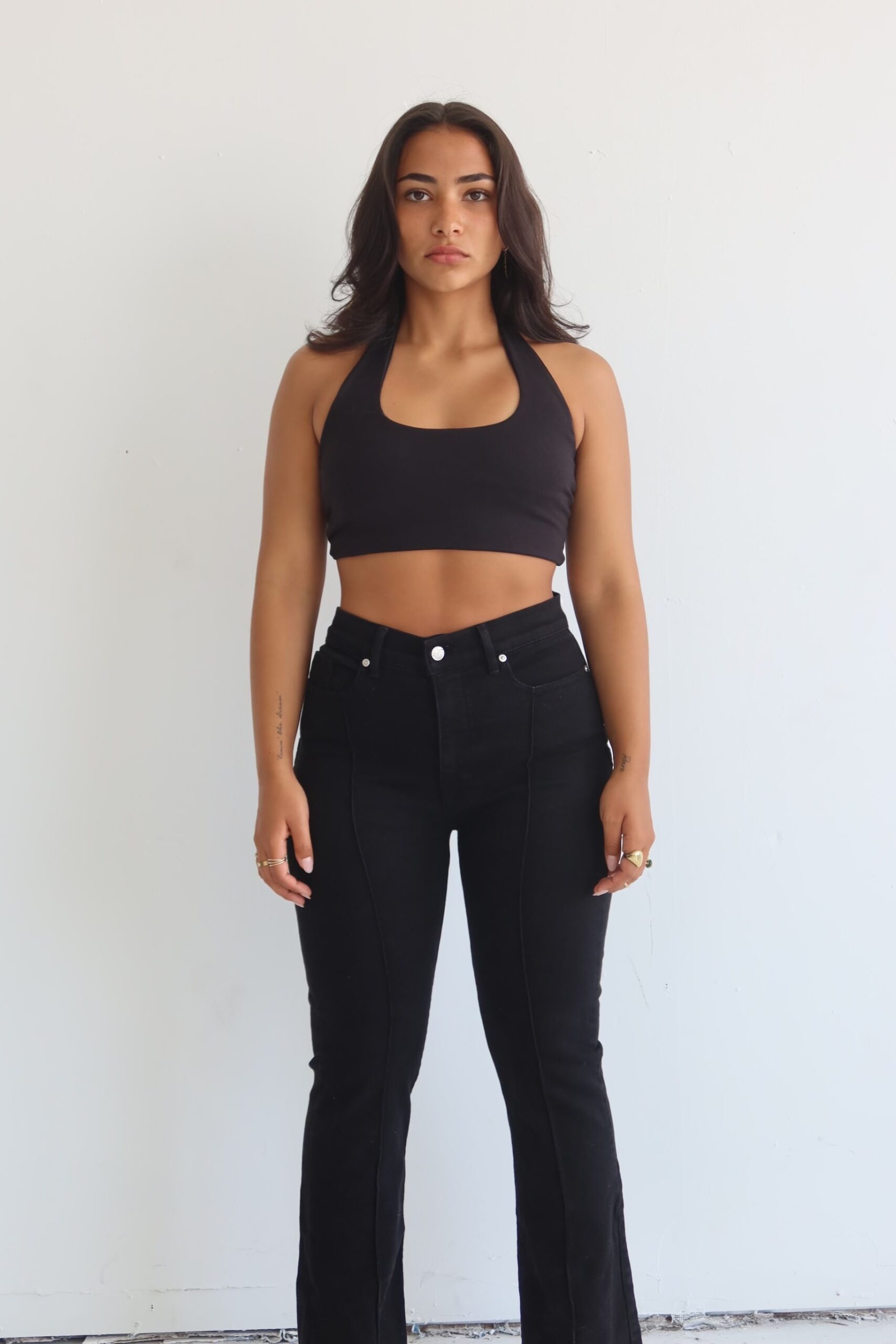 Girl standing against a white backdrop, wearing black jeans and a black halter top with her dark brown hair loosely around her shoulders.