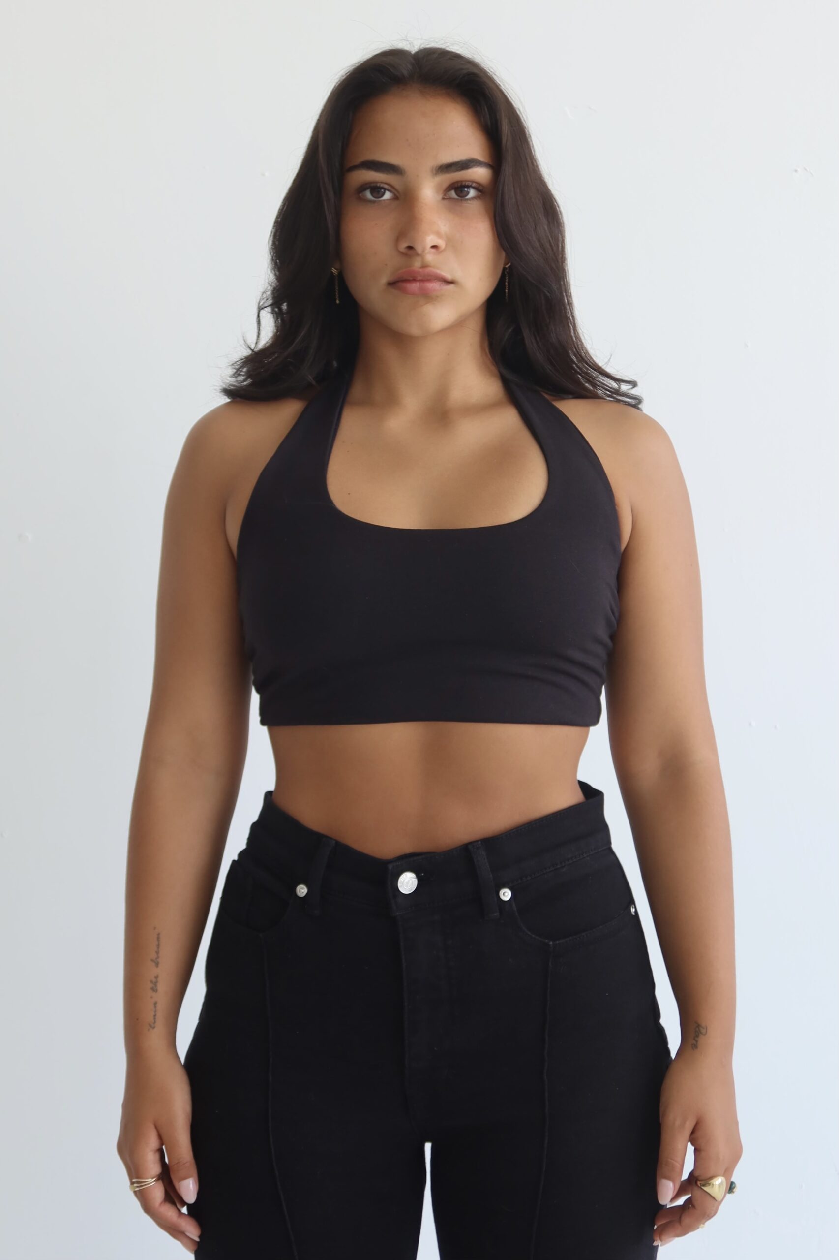 Girl standing against a white backdrop, wearing black jeans and a black halter top with her dark brown hair loosely around her shoulders.