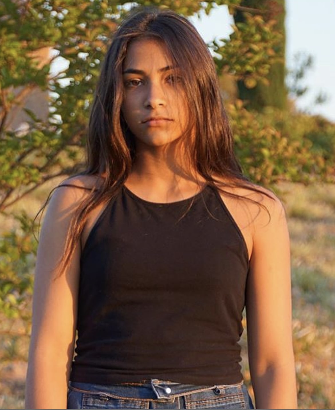 Young girl poses with serious expression in front of a landscape back drop. She wears a black halter top tank top and jeans.