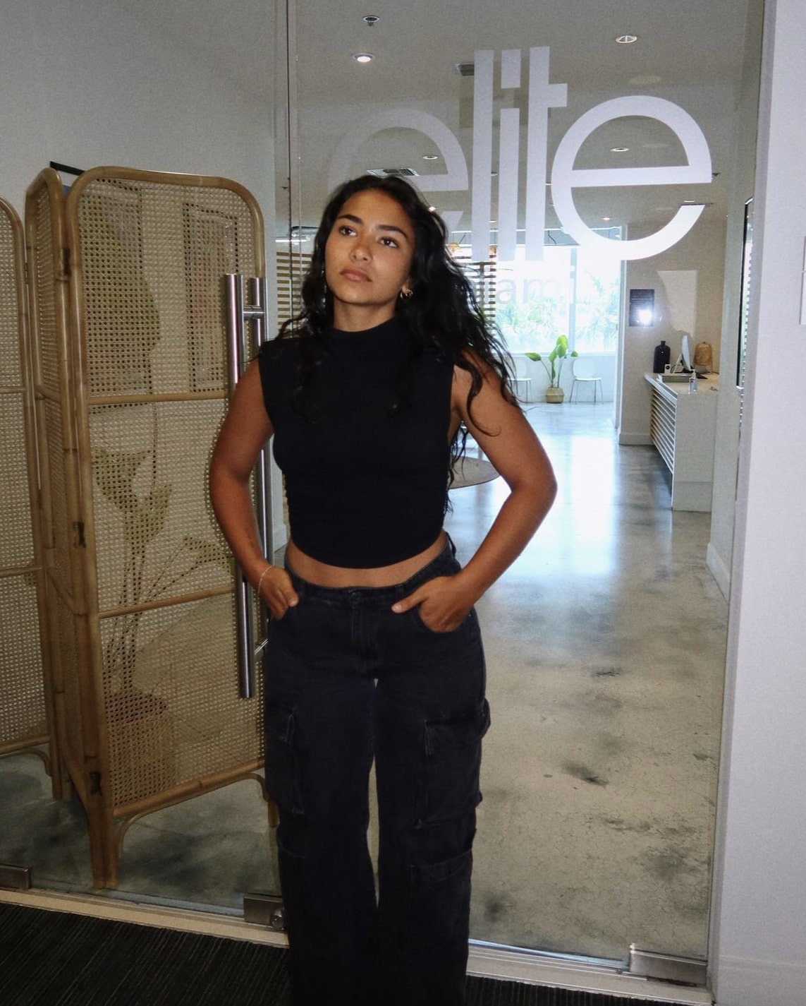 Girl standing in black jeans and black top in front of glass door with opaque text that reads "Elite"