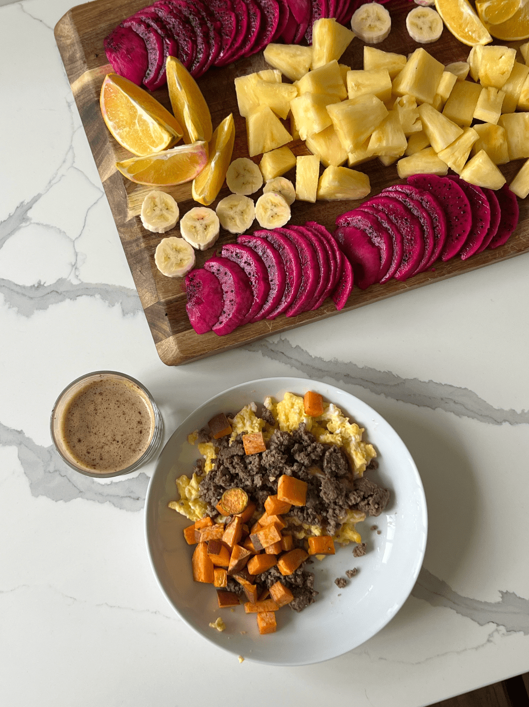 Platter of dragon fruit, bananas, oranges, and pineapple beside a coffee & plate of eggs, sweet potato and ground beef atop a marble counter top