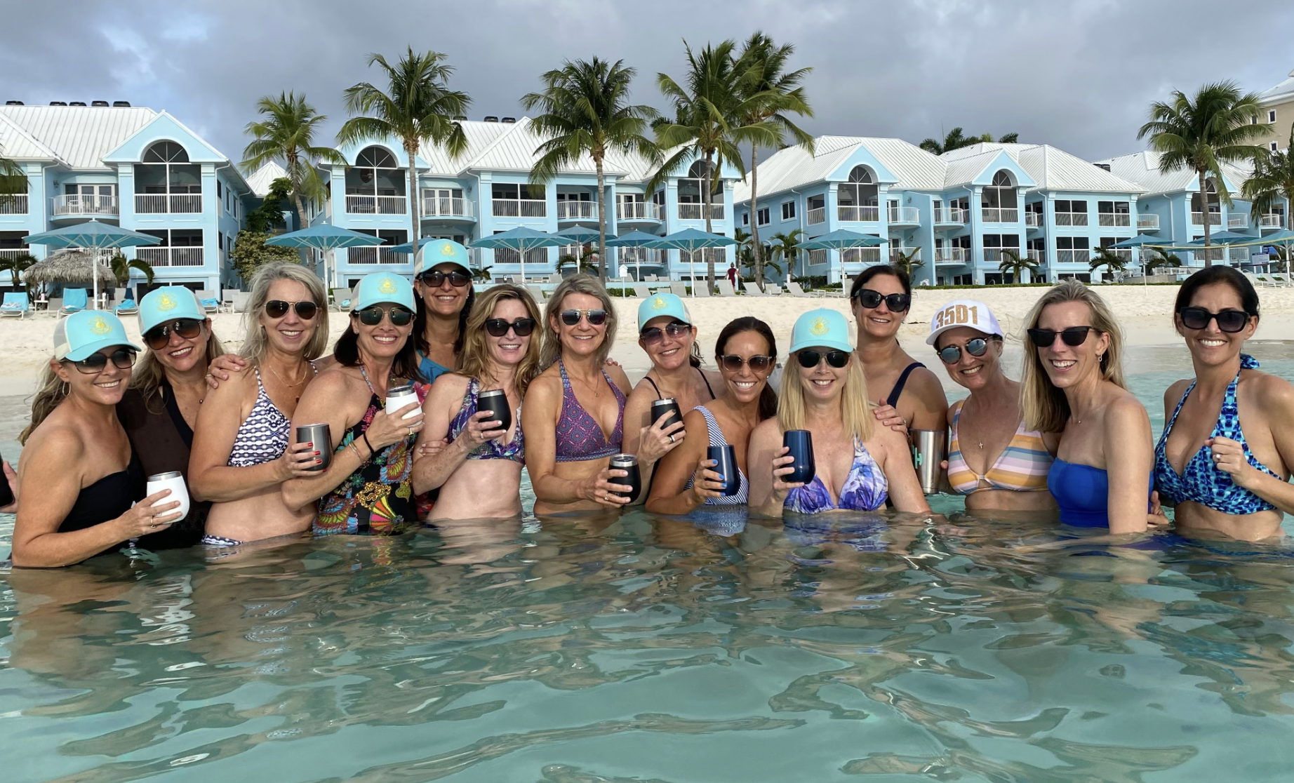 Group of women on annual vacation together, standing in front of a resort waist deep in clear blue water